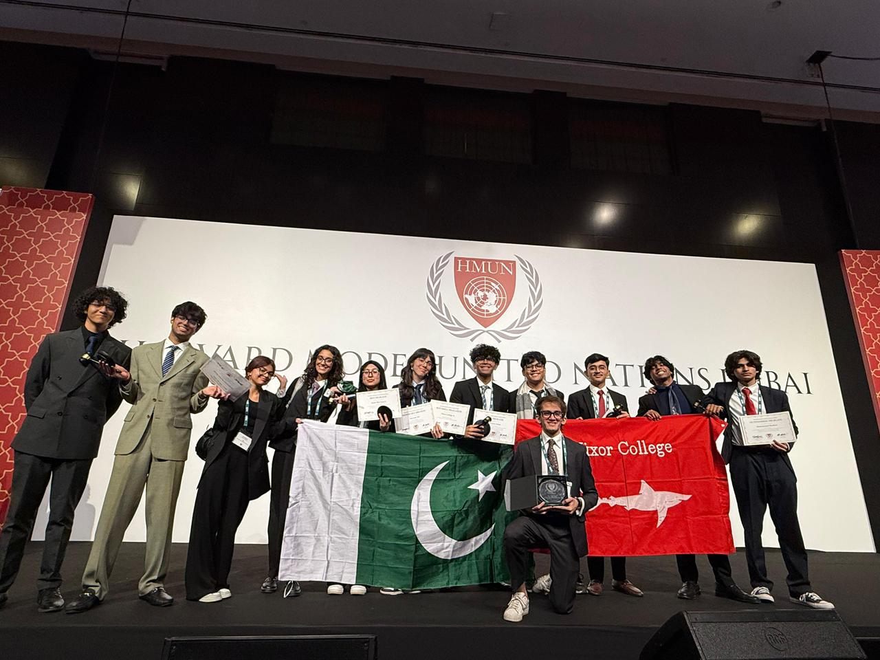 A delegation coached by MUN Society Pakistan on stage at Harvard Model United Nations Dubai, holding the Pakistani flag and award certificates.